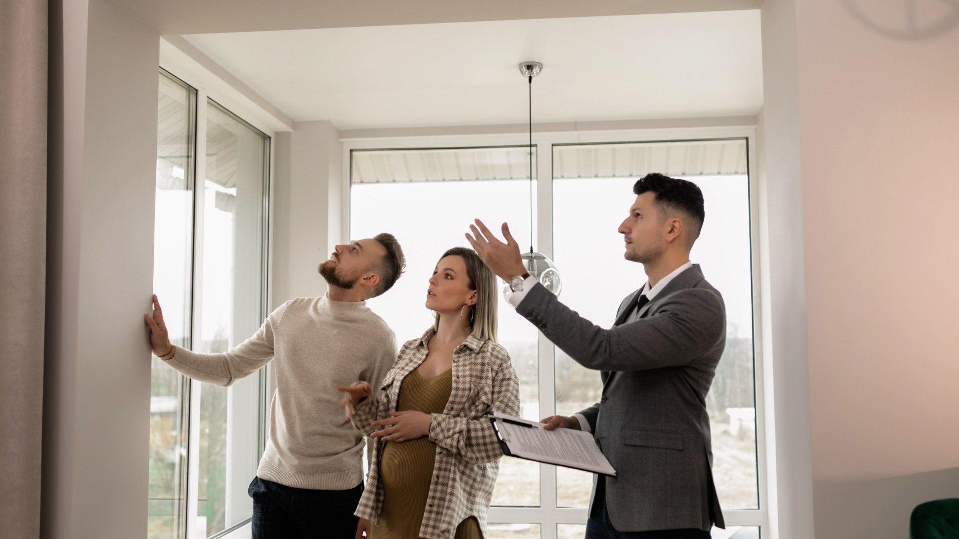 Leasing agent in a gray suit holding a clipboard while gesturing toward the ceiling, showing an apartment unit to a couple standing by a large window