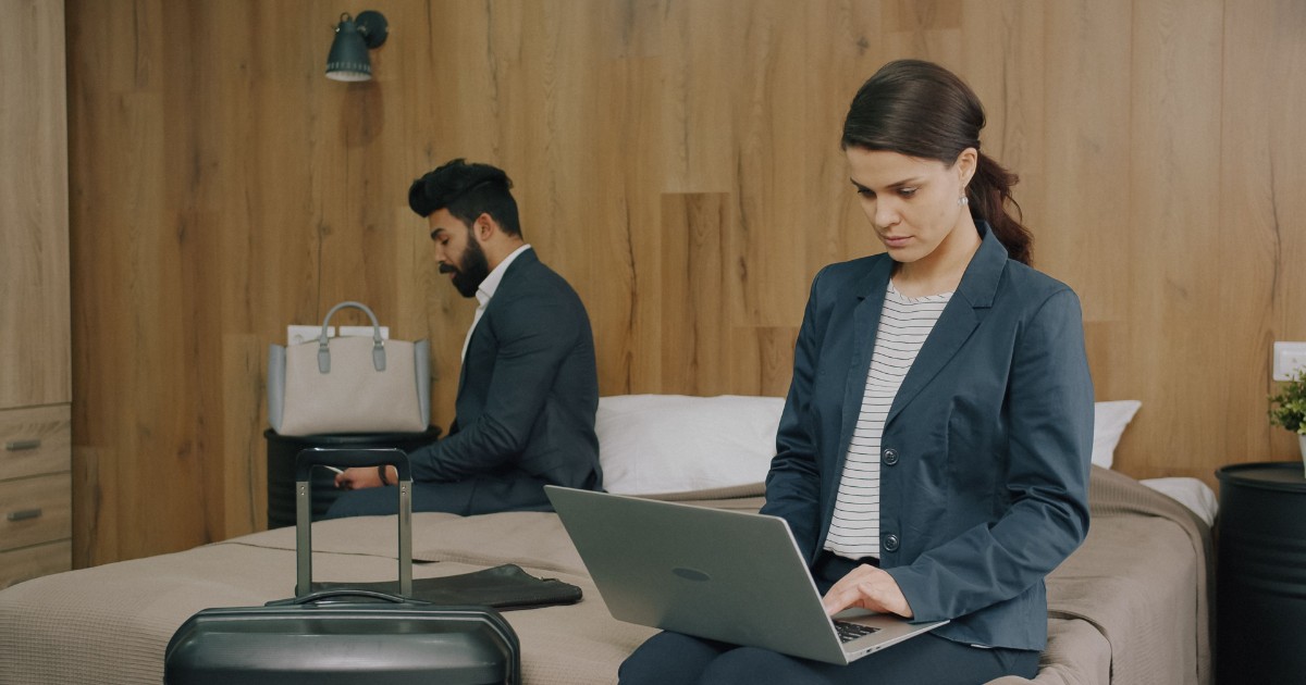 A person works on a laptop in a hotel room while luggage sits nearby, representing temporary housing after an apartment becomes unlivable.