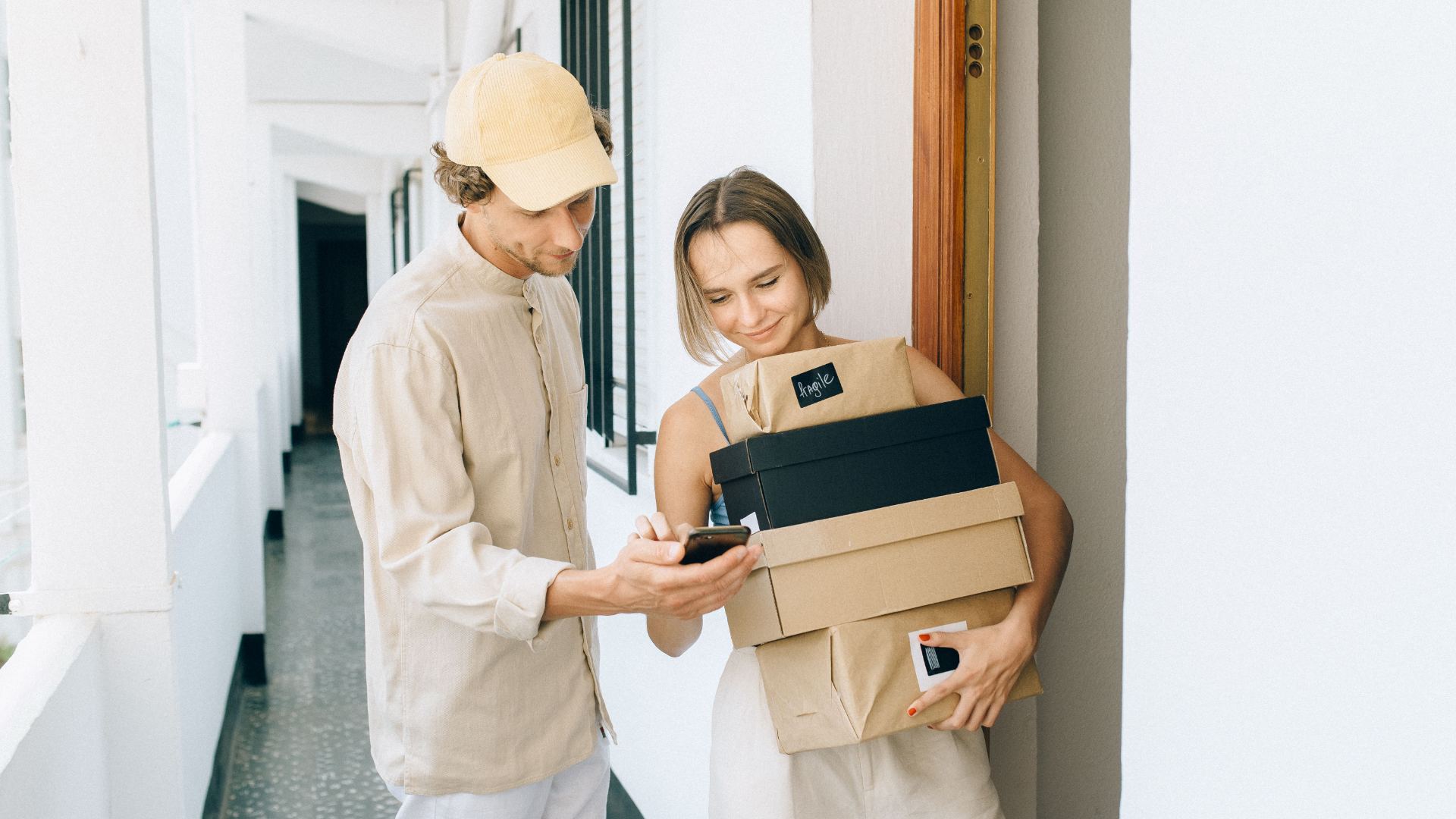 Two apartment residents check a phone while carrying delivered packages in a hallway.