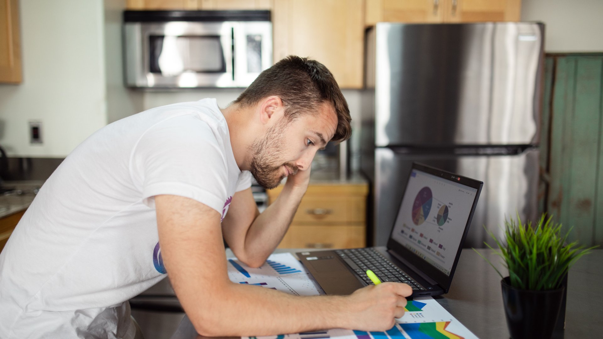 A man reviews financial charts on a laptop and paperwork at his kitchen table.