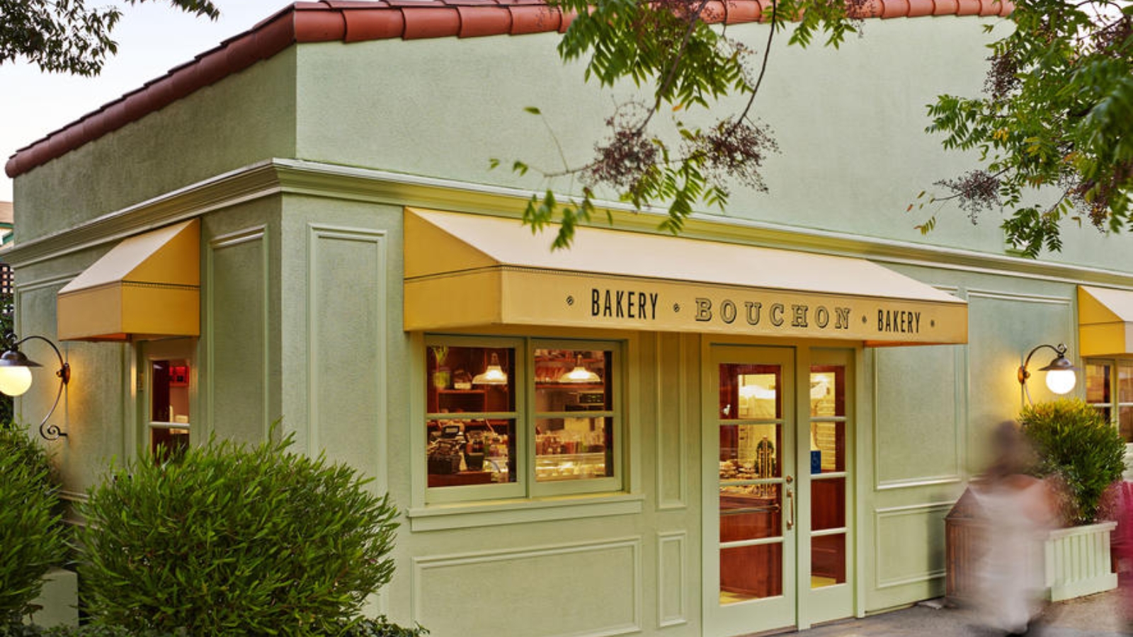 Bouchon Bakery showcases its inviting storefront with distinctive yellow awnings, sage green exterior walls, and warm interior lighting creating a cozy European bakery atmosphere in the evening