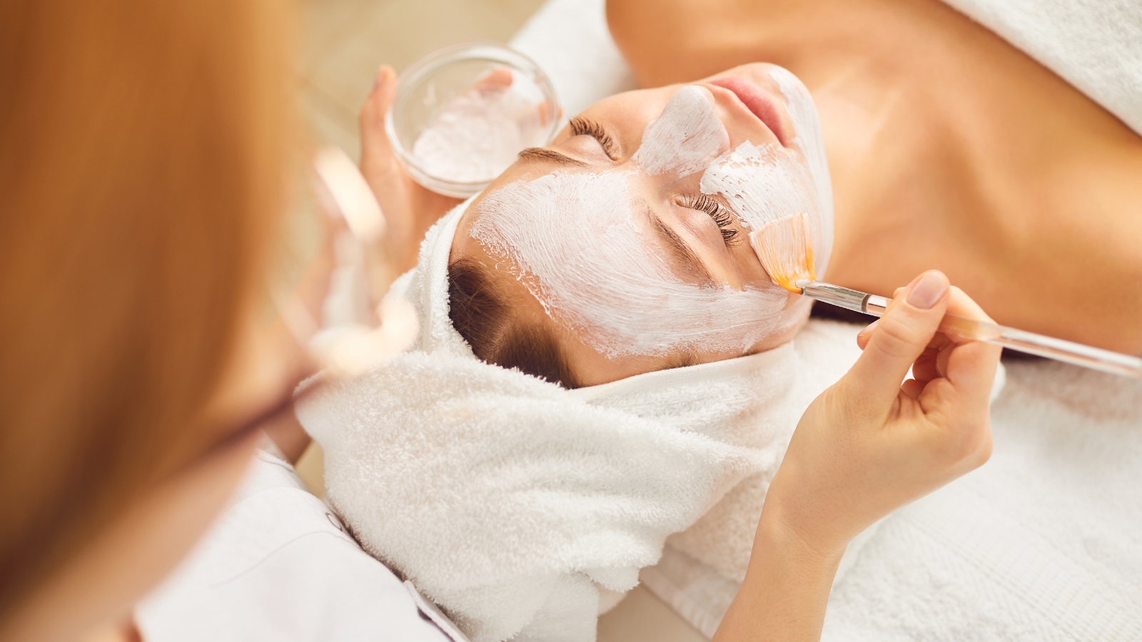Spa Villagio offers luxurious facial treatments with a professional aesthetician carefully applying a rejuvenating white mask to a client's face using a precision brush in a serene spa setting