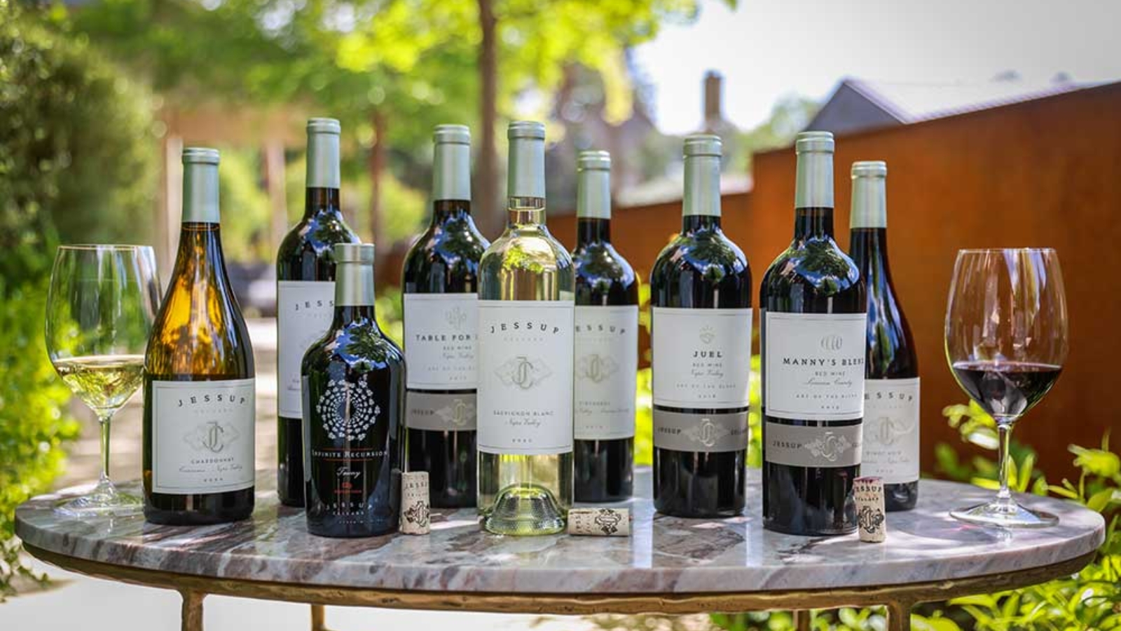 Jessup Cellars presents an outdoor wine tasting featuring multiple bottles of their premium wines arranged on a marble table with filled wine glasses, set against a backdrop of lush greenery and winery buildings