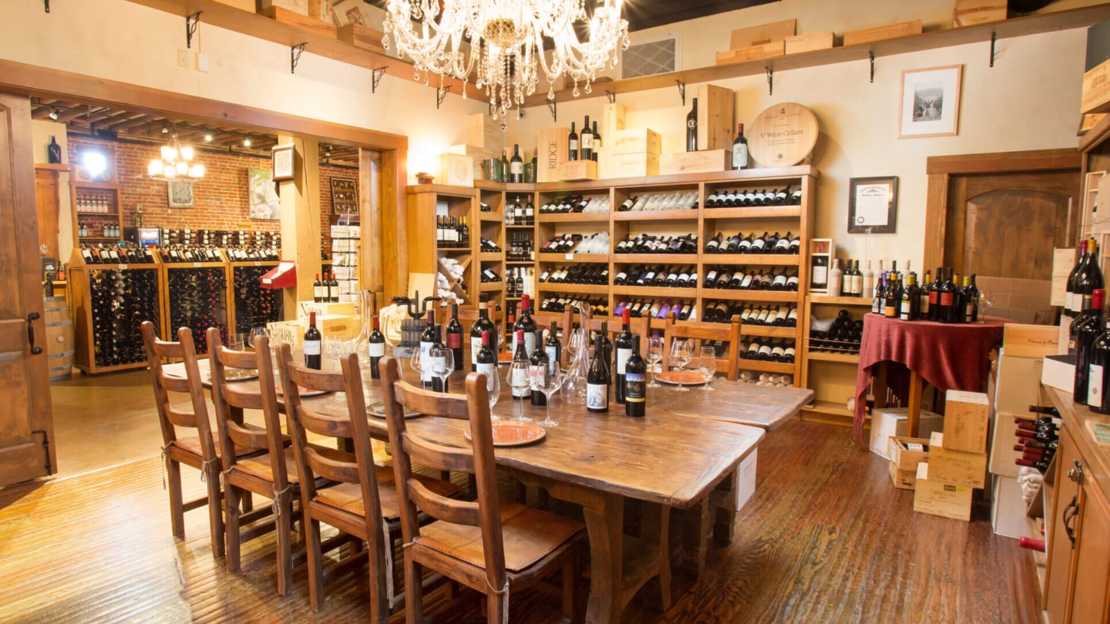 V Marketplace features an elegant wine tasting room with rustic wooden beams, crystal chandelier, extensive wine bottle displays, and a large communal table set up for wine tastings in a warm, welcoming atmosphere