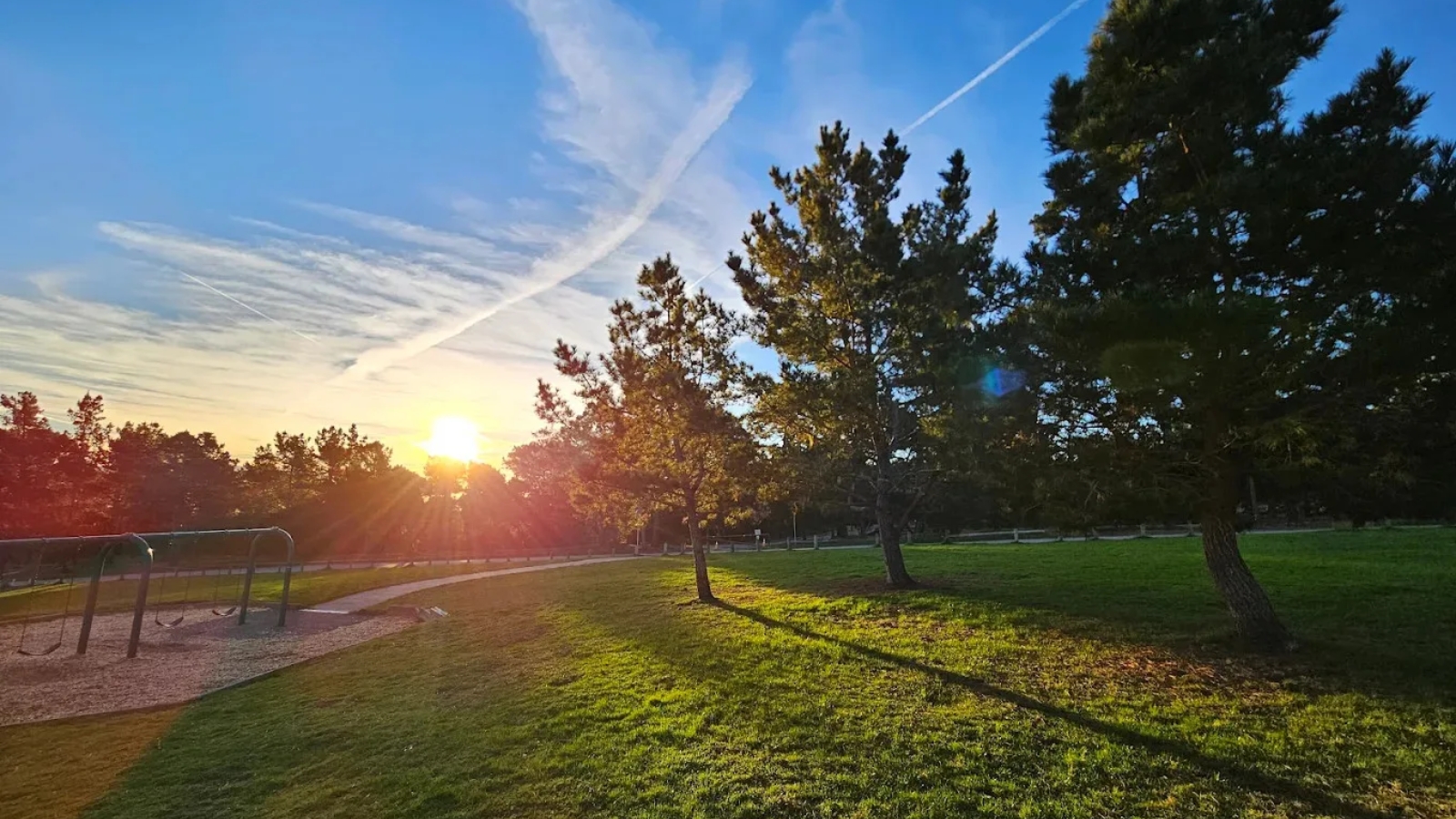 Sunset at Veterans Memorial Park creates a stunning golden hour scene with warm sunlight filtering through mature trees and casting long shadows across the green lawn and walking paths