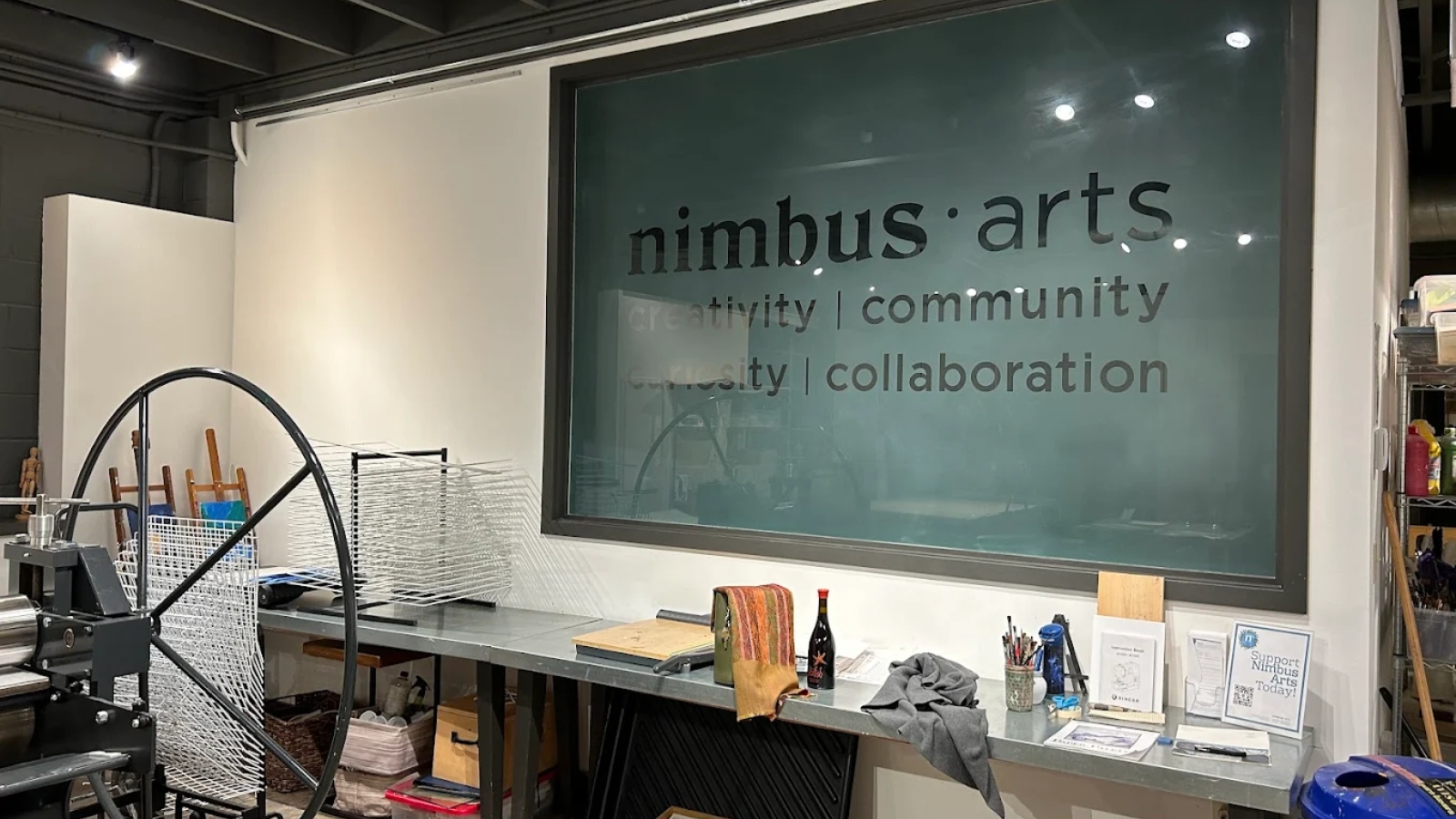 Art studio workspace at the Galleries at Nimbus Arts featuring printmaking equipment, work tables, and educational materials in an industrial setting focused on creativity, community, and collaboration.