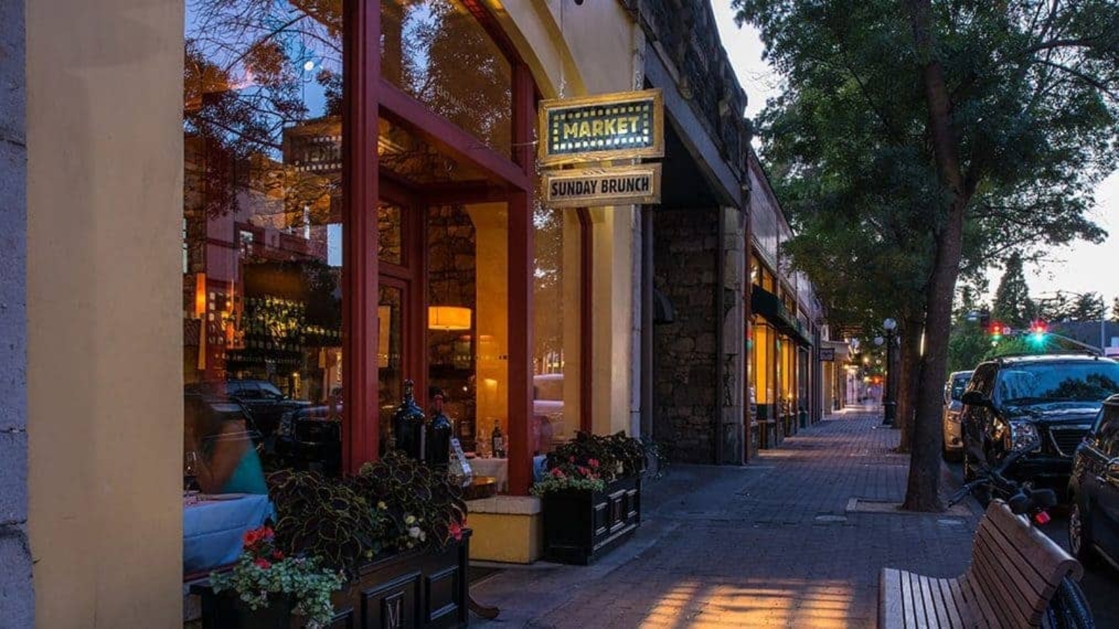 Evening view of Main Street St. Helena featuring illuminated storefronts, sidewalk dining areas with planters, and tree-lined walkways creating a welcoming small-town atmosphere.