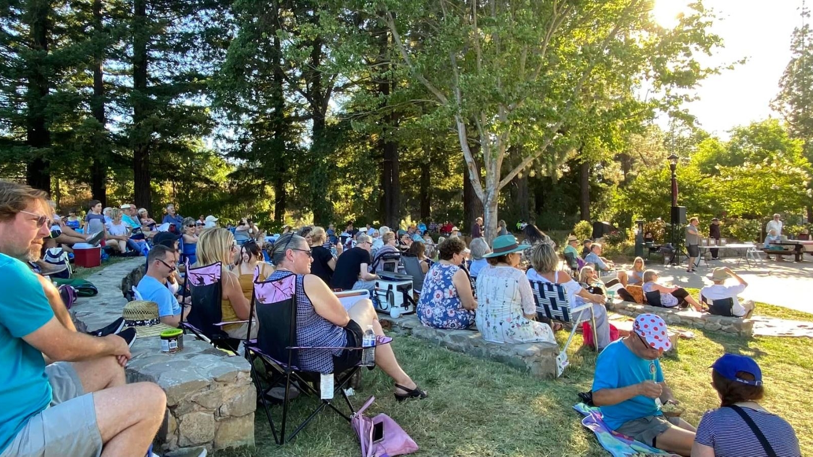 Families and community members gather with blankets and chairs on the grass for Music in the Park Yountville, enjoying an outdoor concert series in the tree-lined park setting during golden hour.