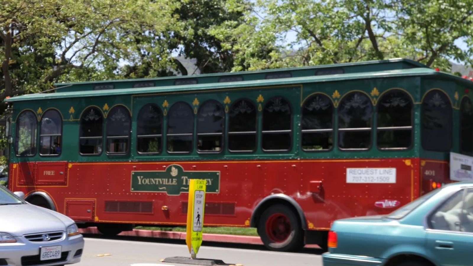 The Yountville Free Trolley, featuring classic green and red vintage-style design with arched windows, travels along a tree-lined street providing complimentary transportation through the Napa Valley town.