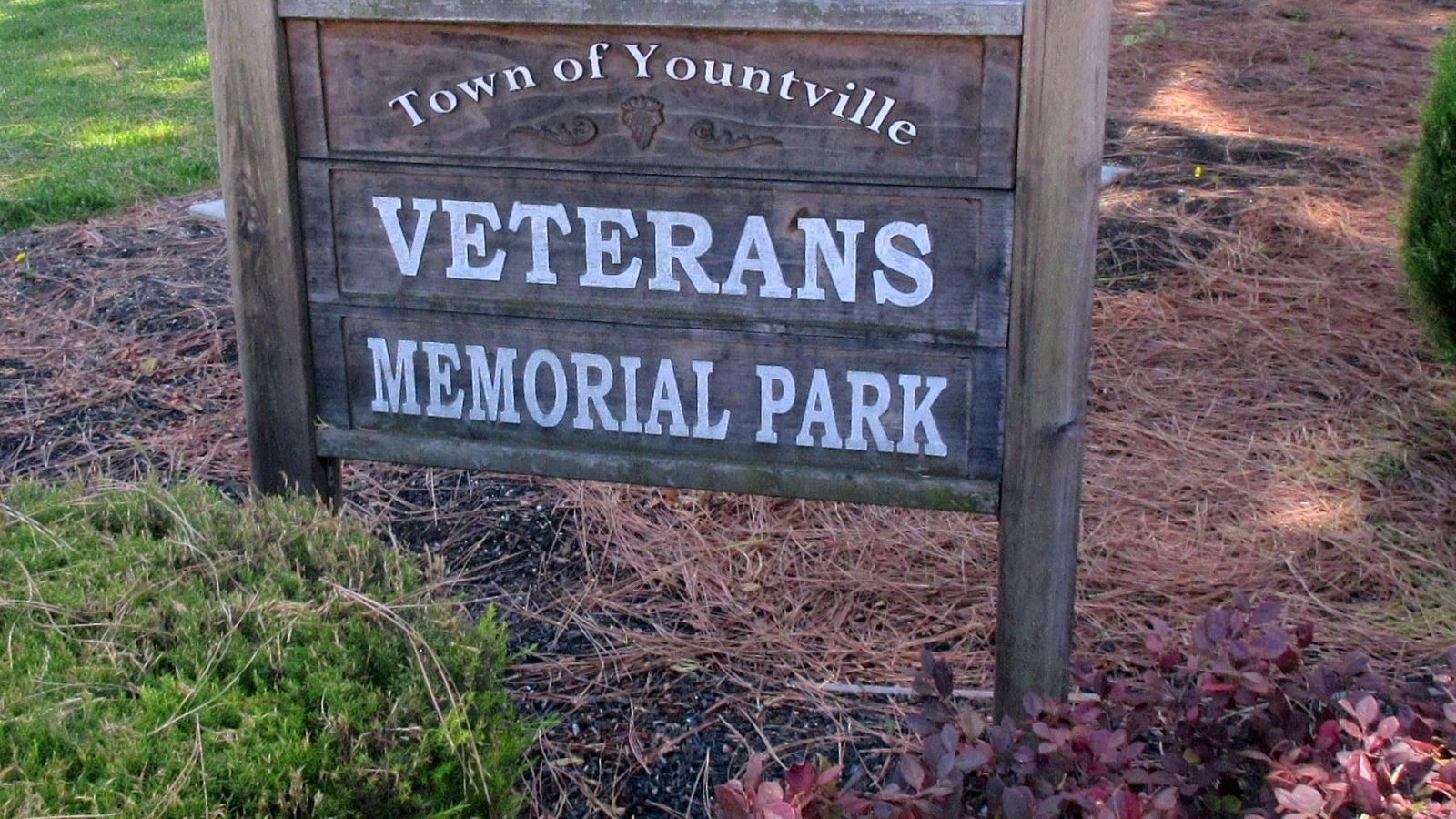 Wooden entrance sign for Yountville Veterans Memorial Park displaying "Town of Yountville" at the top and "Veterans Memorial Park" below, surrounded by landscaped grounds and autumn foliage.