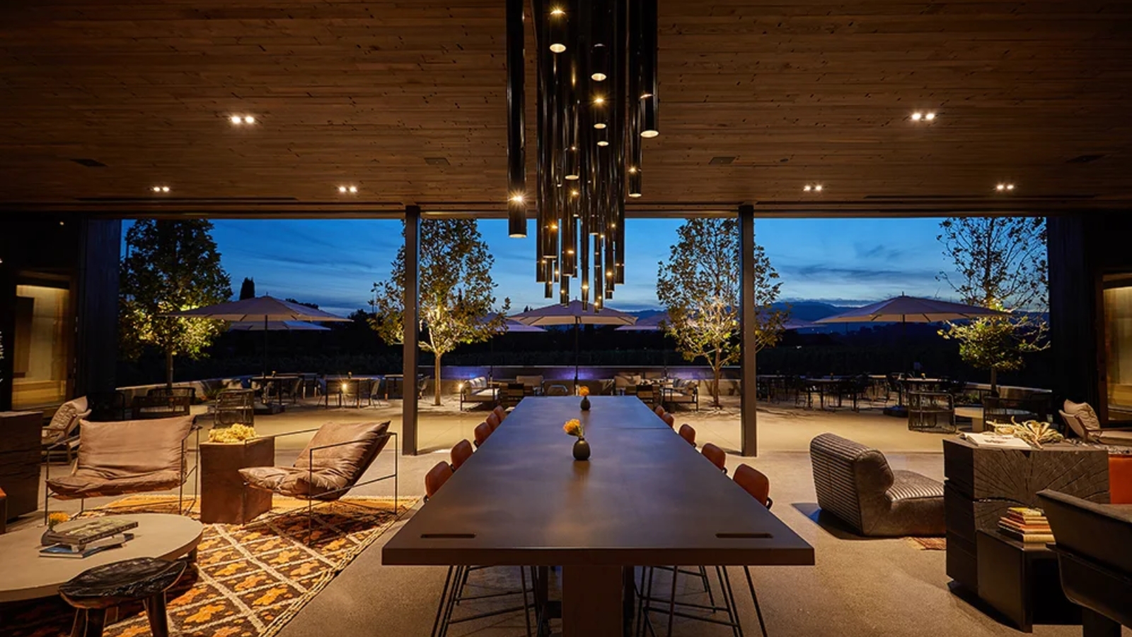 Modern tasting room interior at Clos du Val featuring a long wooden dining table with contemporary seating, dramatic pendant lighting, and floor-to-ceiling windows showcasing panoramic vineyard views during twilight hours.