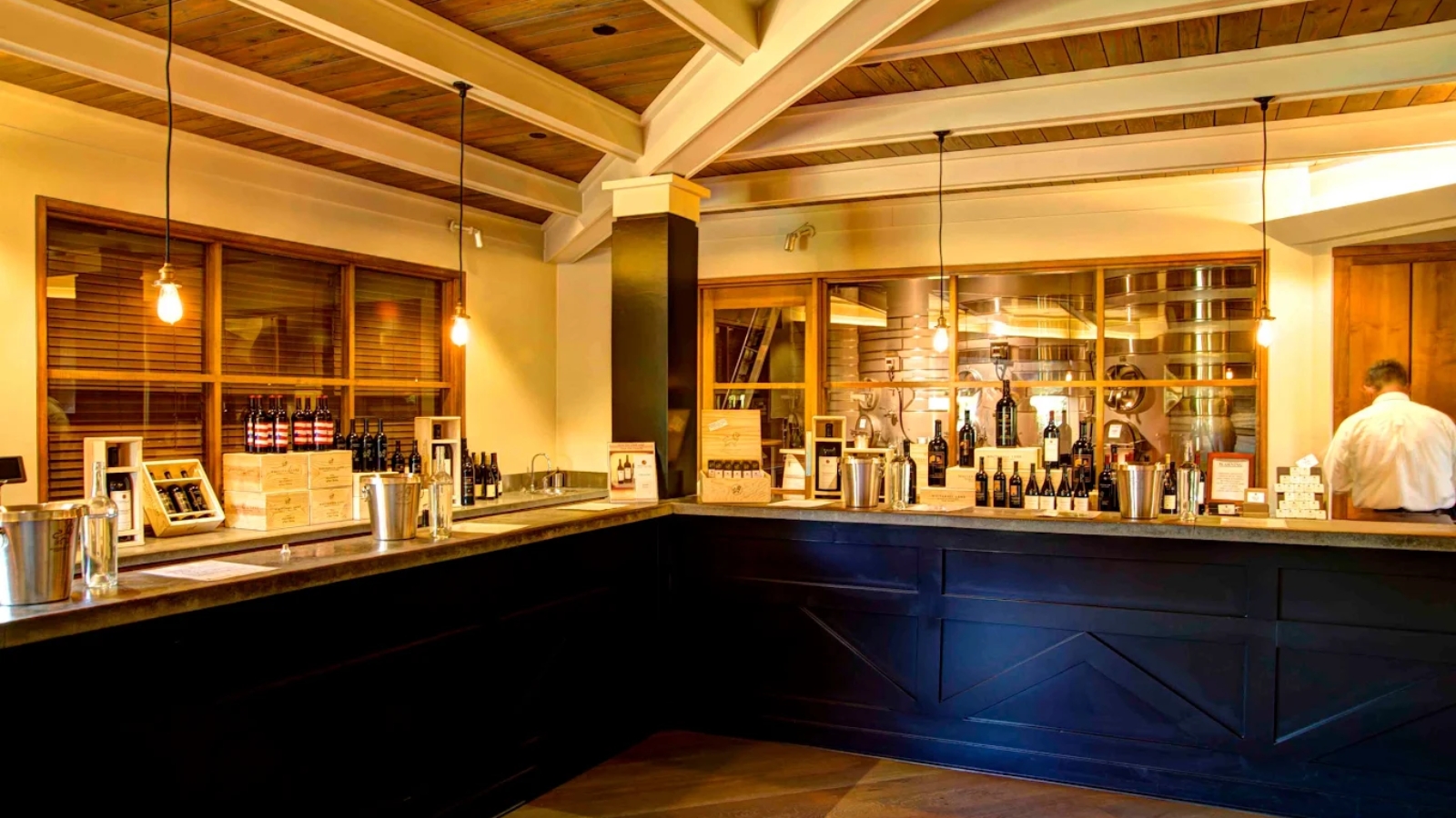 Elegant tasting room interior at Whitehall Lane Winery featuring exposed beam ceiling, dark blue cabinetry, extensive wine bottle displays, and warm pendant lighting creating a sophisticated wine tasting environment with rustic charm.