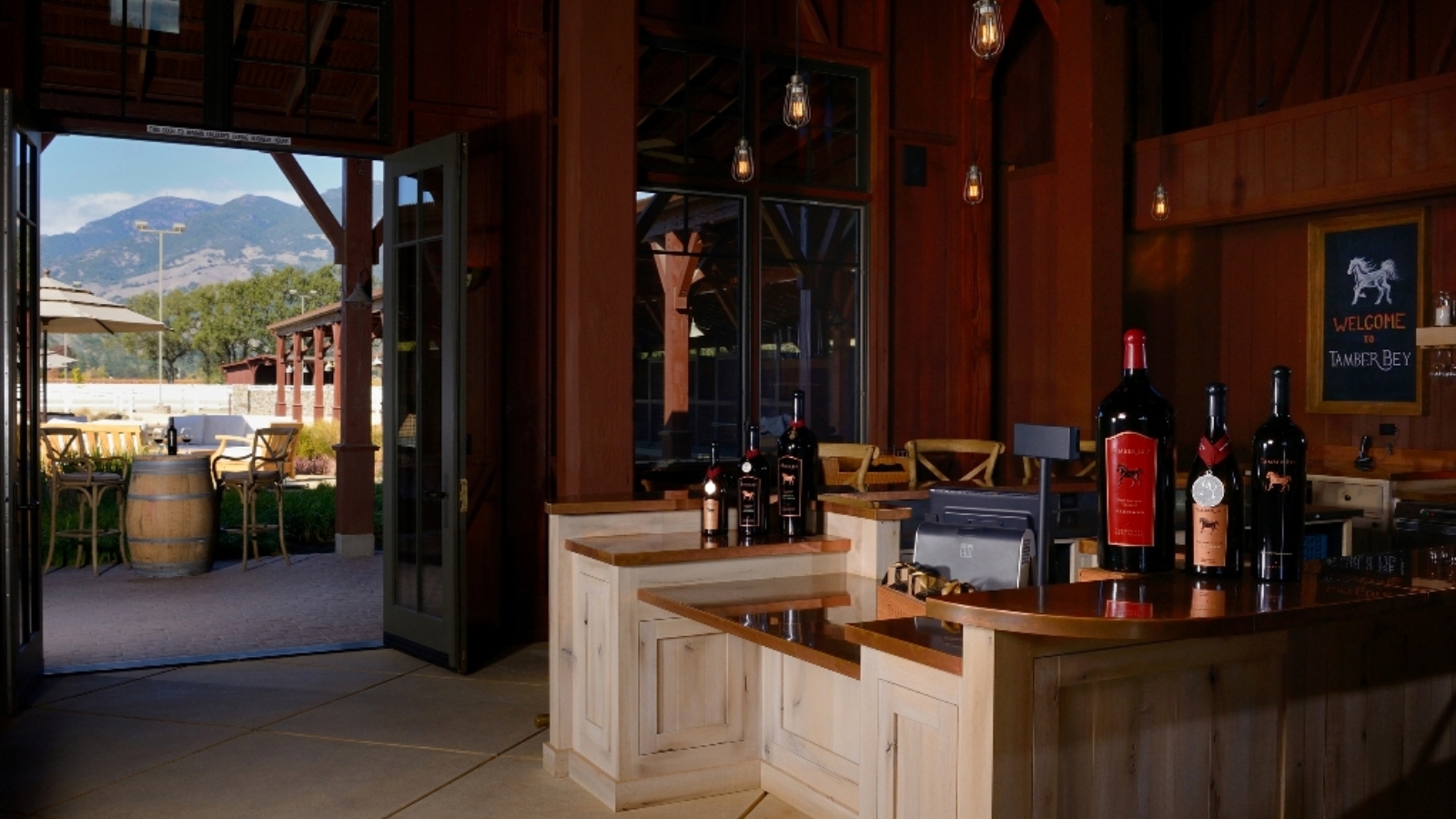 Rustic tasting room interior at Tamber Bey Vineyards featuring industrial lighting, exposed wooden beams, a white wooden tasting counter with wine displays, and an open doorway revealing mountain views and outdoor patio seating.