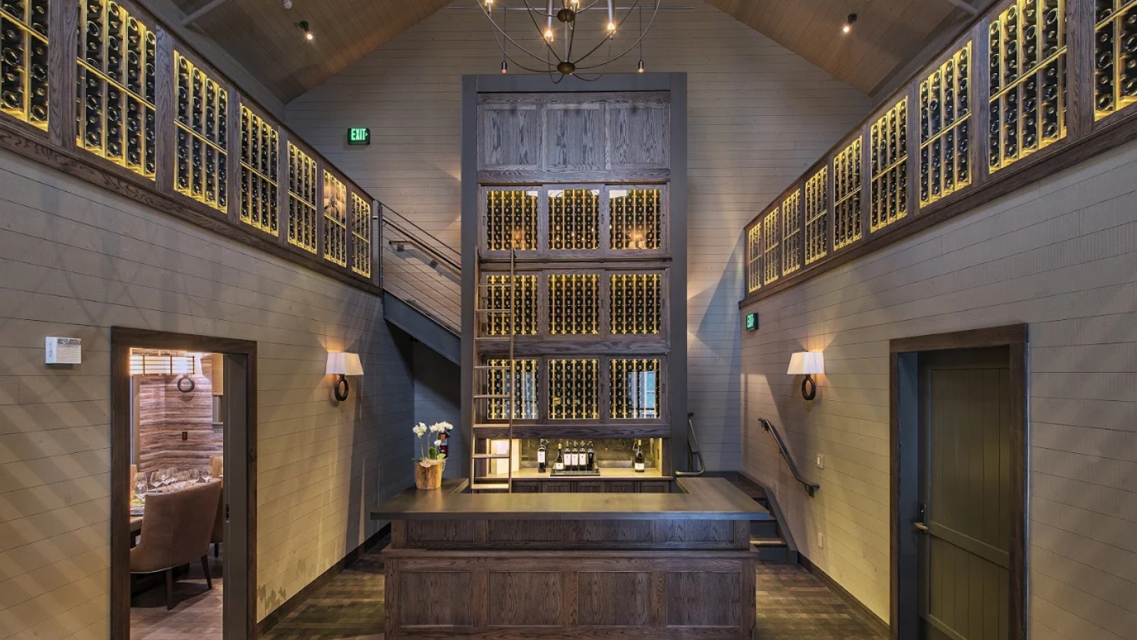Spectacular multi-level wine cellar at Alpha Omega Winery featuring floor-to-ceiling illuminated wine storage, a central wooden tasting bar, dramatic chandelier lighting, and sophisticated modern architecture creating an impressive wine library atmosphere.