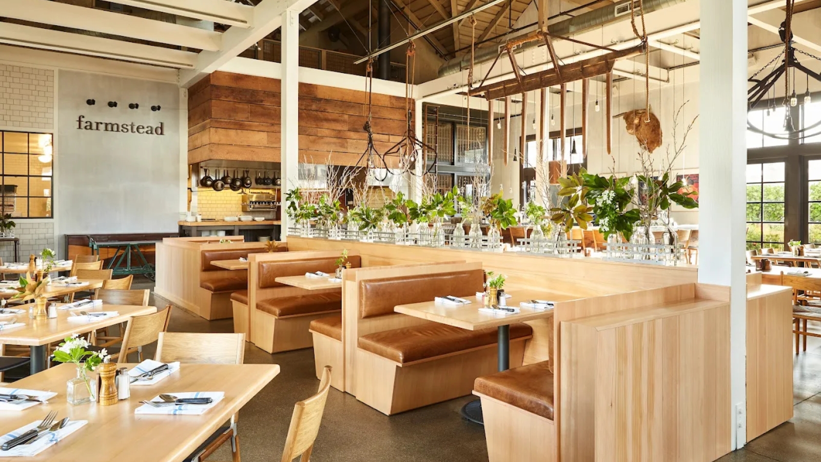 Modern rustic interior of Farmstead At Long Meadow Ranch restaurant featuring exposed beam ceilings, leather banquettes, natural wood furnishings, and a central bar adorned with fresh greenery.