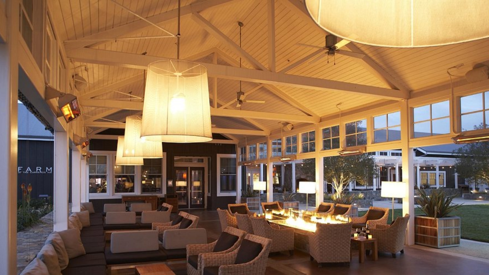 Elegant covered outdoor lounge at The Farm At Carneros Resort featuring wicker seating around fire tables, pendant lanterns, and exposed beam ceilings overlooking the resort grounds at dusk.