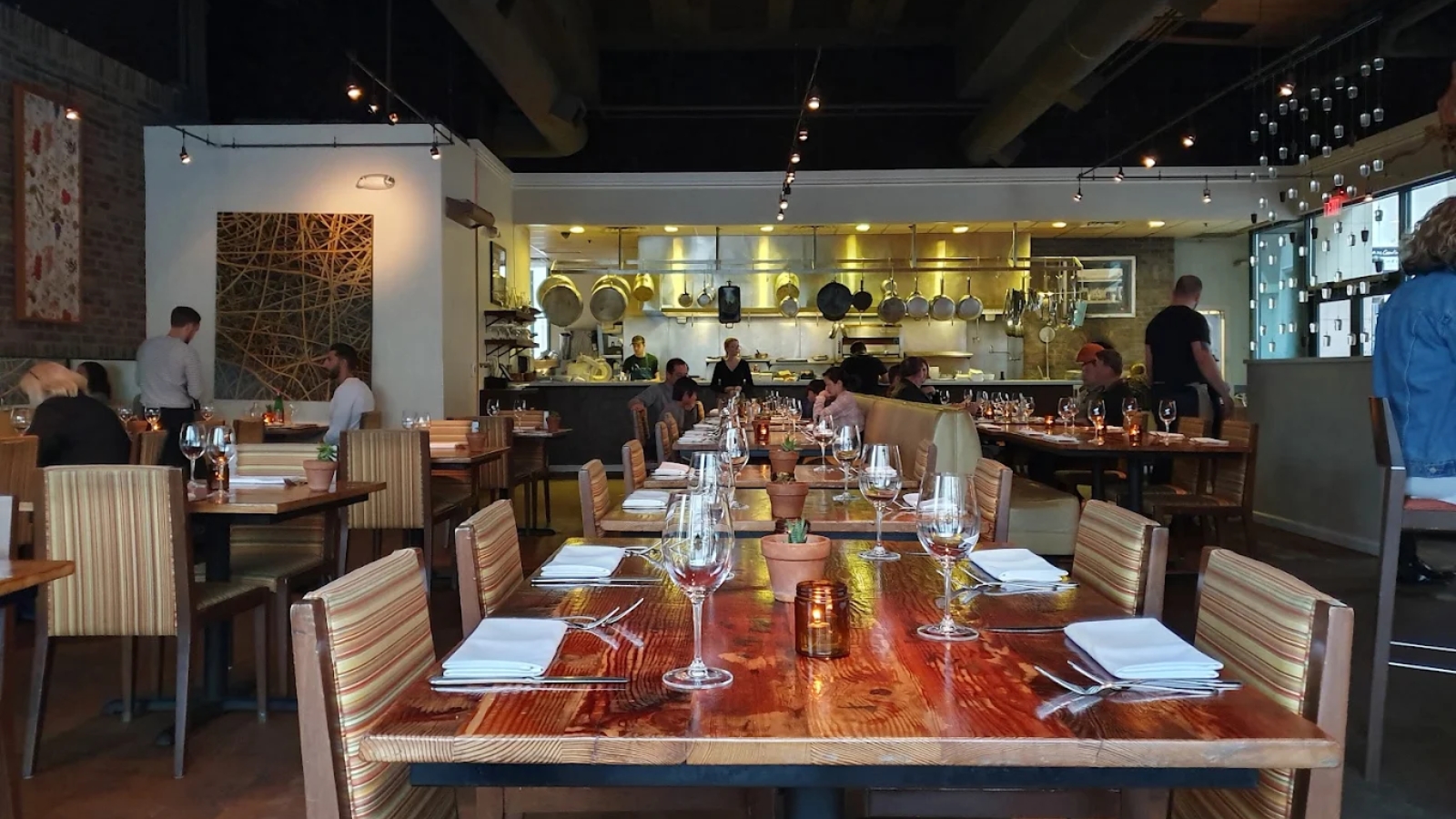 Modern Italian restaurant interior at Oenotri featuring an open kitchen, wooden communal tables set for dinner, and contemporary track lighting with exposed ceiling beams.