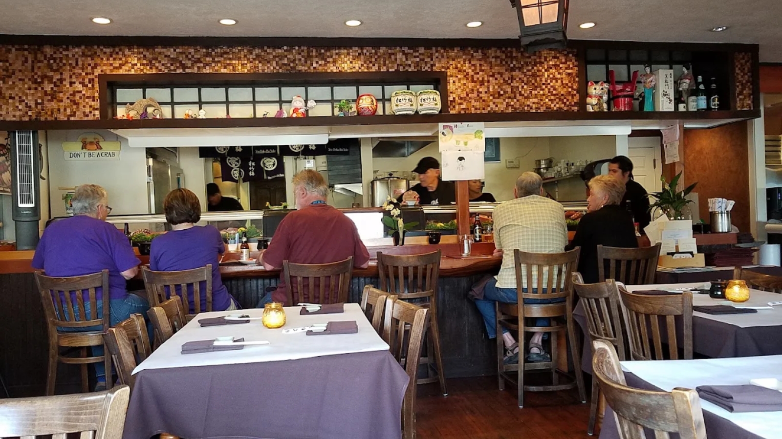 Sushi Mambo restaurant interior featuring wooden counter seating with customers, brick mosaic accent wall, paper lantern lighting, purple tablecloths, wooden chairs, and open kitchen with staff visible.