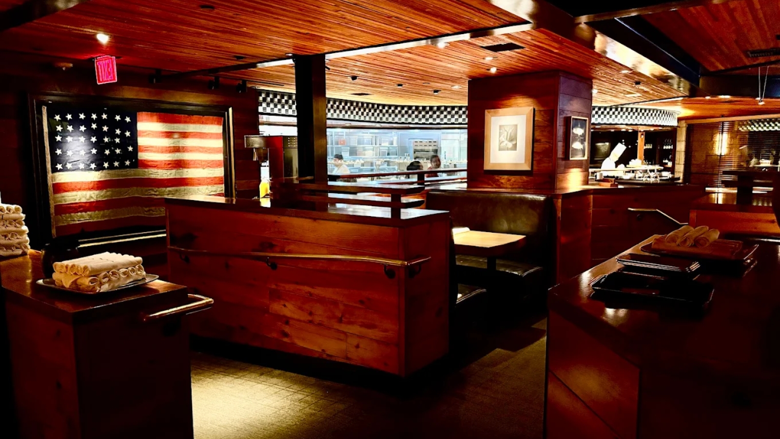 R+D Kitchen restaurant interior featuring warm red wood architecture, American flag artwork, checkered pattern accents, wooden counter seating with displayed dishes, warm ambient lighting, and modern culinary design.