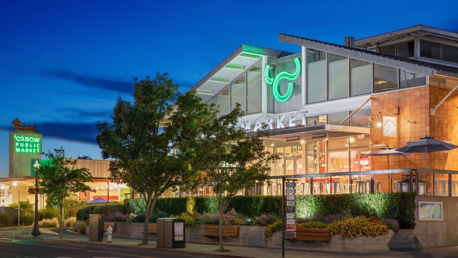 Oxbow Public Market illuminates the evening with neon signage and architectural lighting, showcasing the modern marketplace entrance with landscaped planters and outdoor seating area.