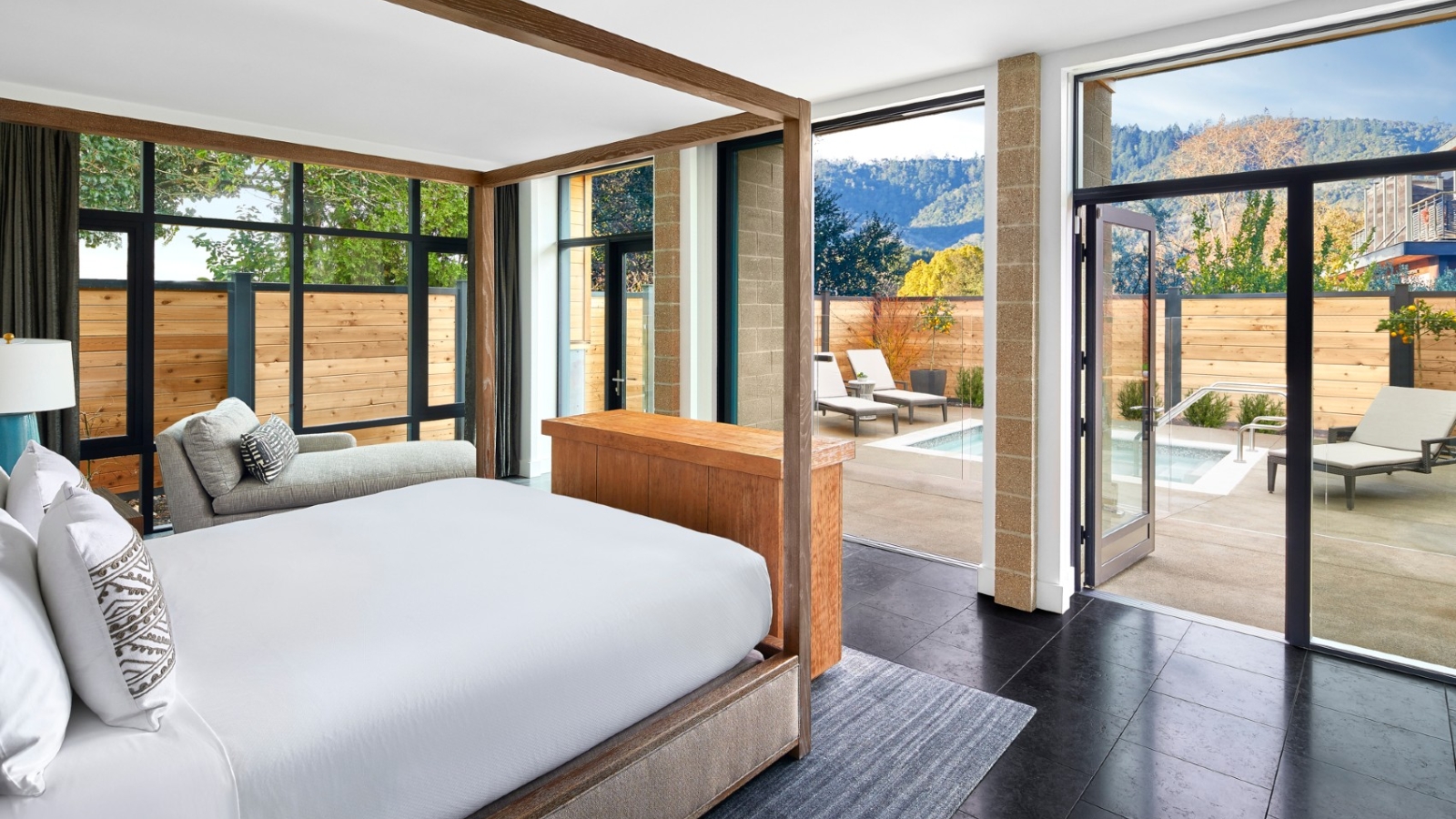 A luxury suite at Bardessono Hotel & Spa features a wooden four-poster bed with white linens, floor-to-ceiling windows framing vineyard and mountain views, a private patio with lounge seating, and contemporary furnishings.