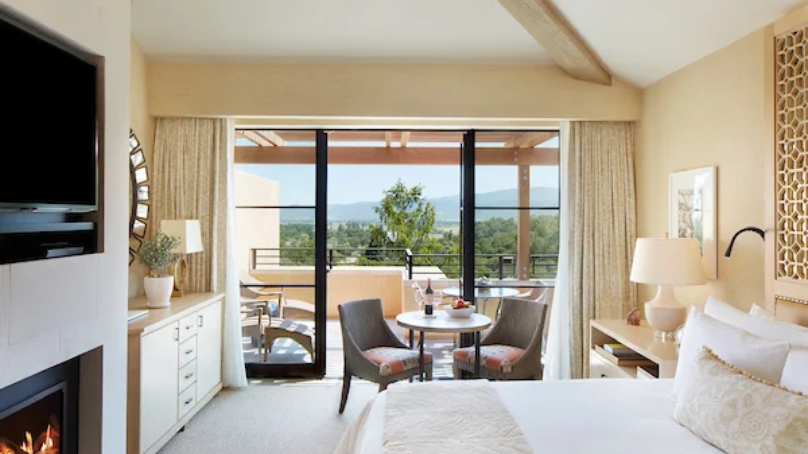 A luxurious guest suite at Auberge du Soleil showcases a king bed with cream bedding, floor-to-ceiling windows with vineyard valley views, a fireplace, and a private balcony with seating overlooking the landscape.