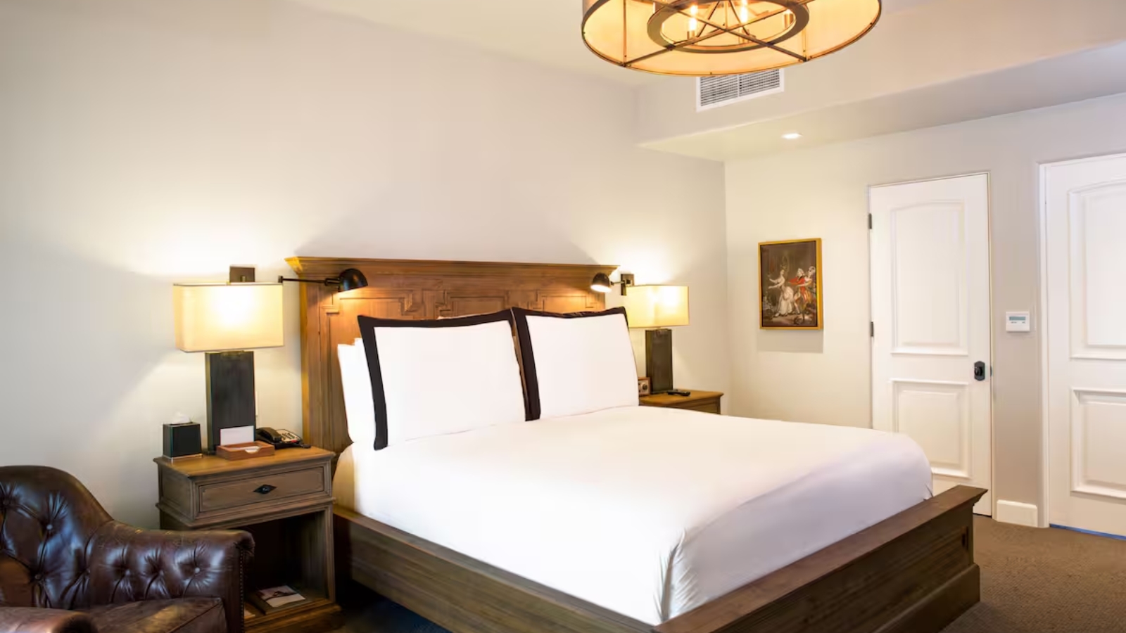 An elegantly appointed guest room at North Block Hotel features a king bed with white linens and black accent pillows, warm ambient lighting from table lamps and a modern pendant fixture, and classic furnishings.