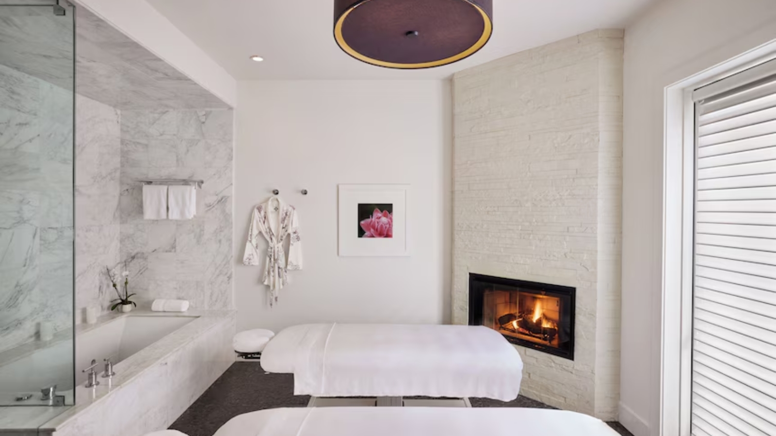 Bardessono Hotel & Spa's luxurious spa treatment room features a massage table, marble-accented walls, a fireplace, and elegant furnishings with a circular ceiling fixture