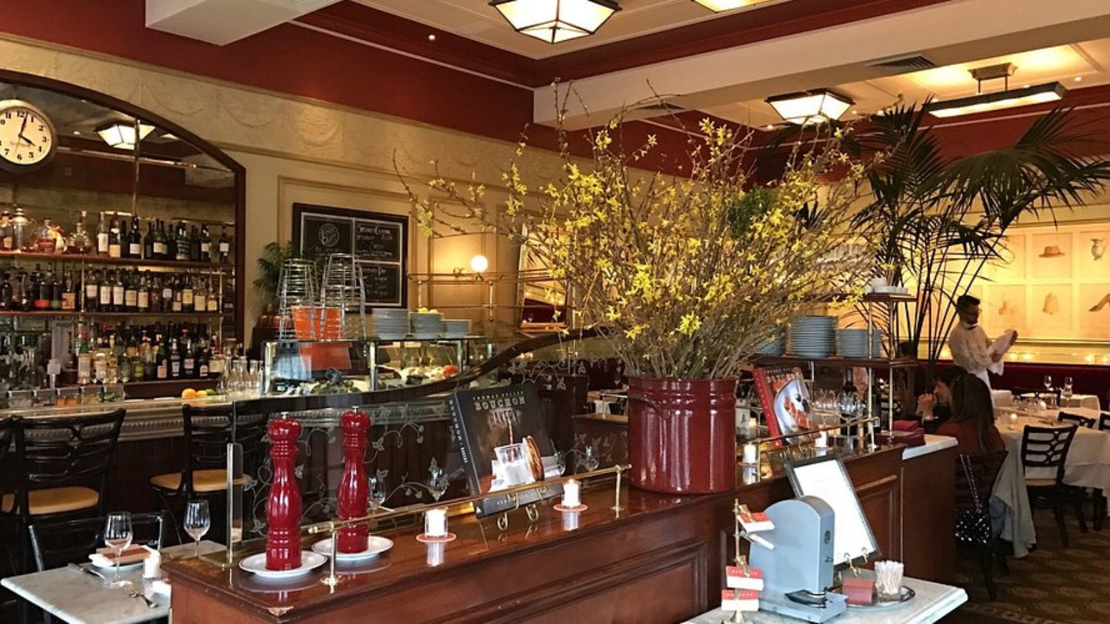 Bouchon Bistro's elegant bar and dining area features warm wood tones, decorative floral arrangements, and a well-stocked bar with wine bottles and glassware.