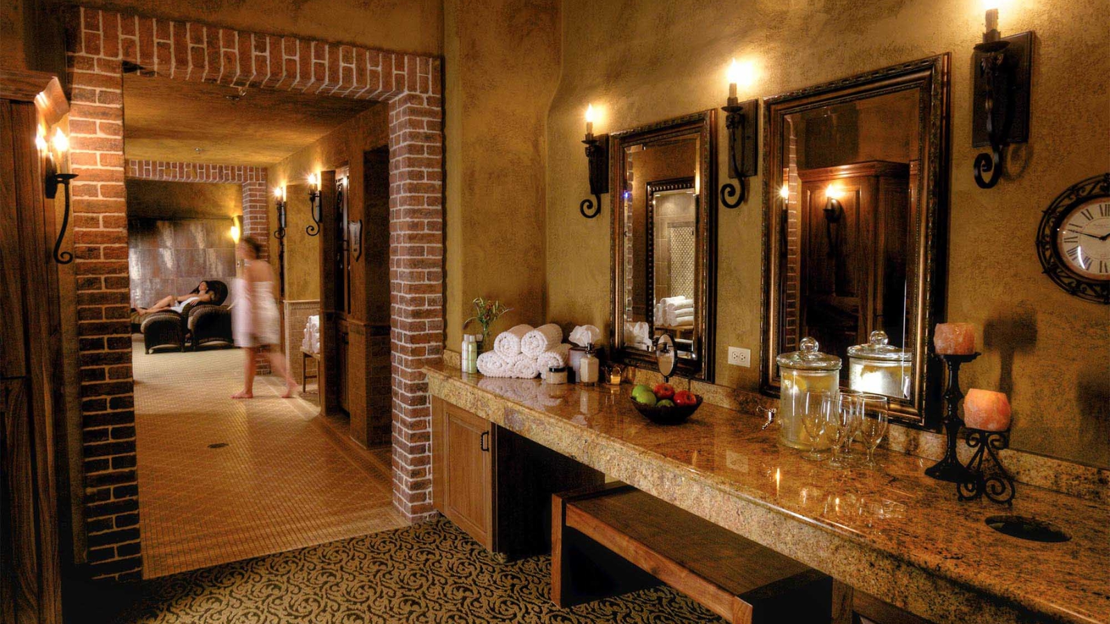 Spa Terra At The Meritage Resort features a luxurious spa bathroom with dual vanities, ornate mirrors, warm lighting, rolled towels, and access to a brick fireplace relaxation area.