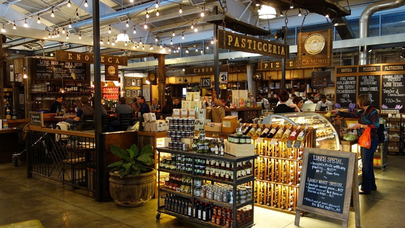 Oxbow Public Market features artisanal food vendors including a pasticceria with specialty products, prepared foods, and gourmet items displayed under warm lighting in a bustling marketplace atmosphere