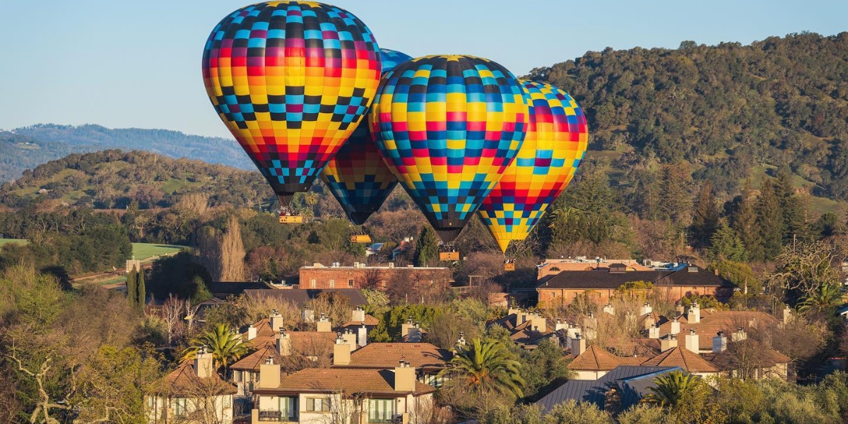 Nava Valley Balloons sunrise balloon flight featuring three colorful hot air balloons with blue, red, yellow, and pink checkered patterns floating above a picturesque European village surrounded by rolling forested hills.