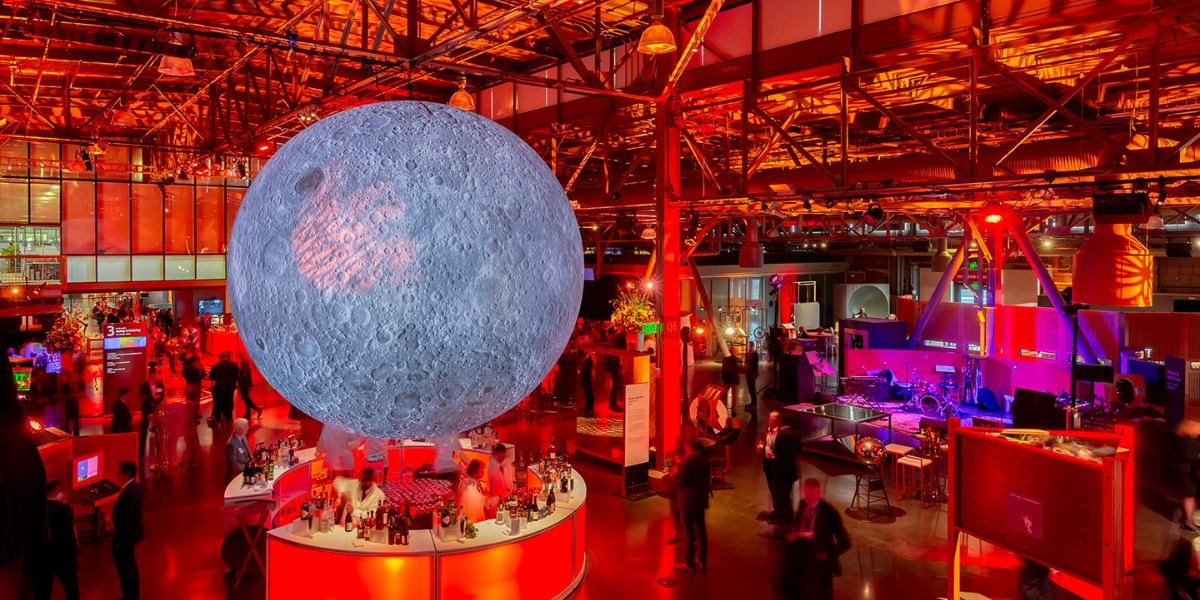 Exploratorium interactive exhibit featuring a massive illuminated moon installation suspended above visitors in a warehouse gallery with vibrant red and purple ambient lighting.