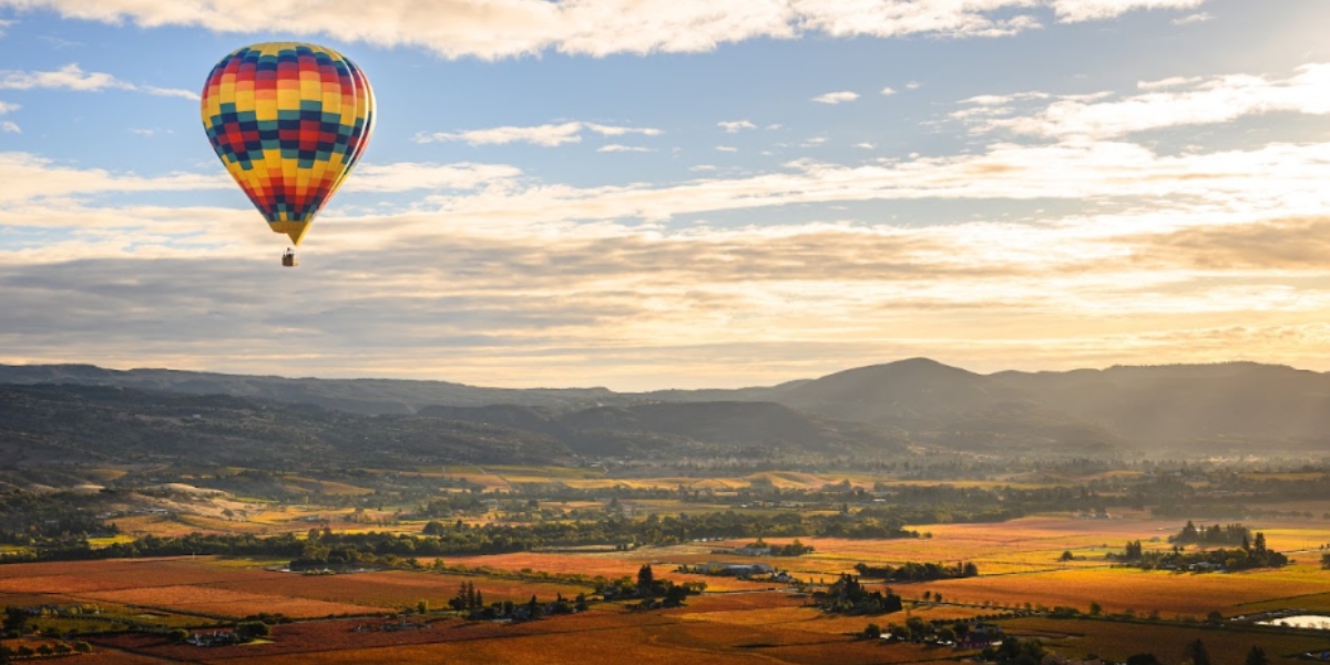 Sunrise balloon flight Napa Valley balloons featuring a colorful hot air balloon floating above rolling vineyard landscapes, golden fields, and mountain ranges during early morning light.