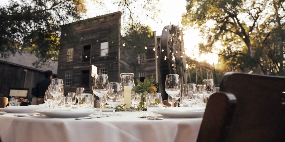 Harvest Dining featuring an elegant outdoor table setting with wine glasses, white linens, and floral centerpieces overlooking historic winery buildings and oak trees at sunset.