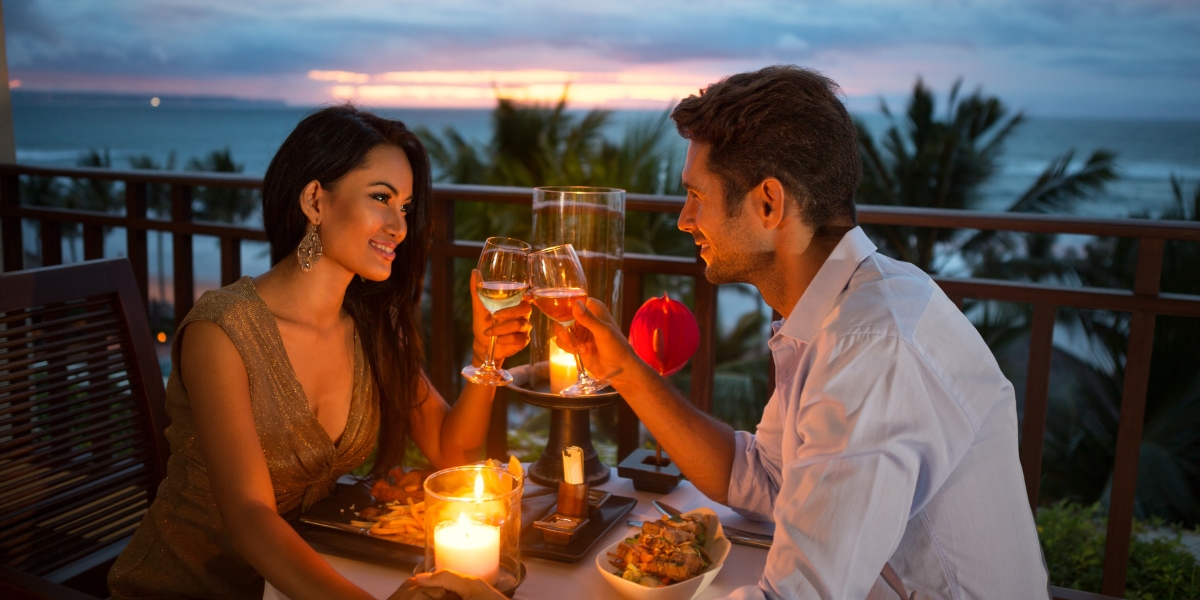 Weekend trips - Celebrate With a Romantic Dinner – couple toasting wine glasses at candlelit oceanside table with sunset sky, palm trees, and romantic ambiance during evening celebration
