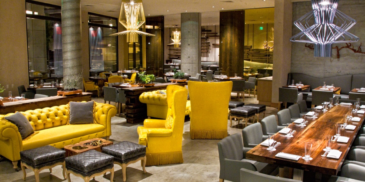 A vibrant dining lounge with bold yellow velvet furniture, sculptural chandeliers, and communal wood tables reflecting the striking interior design of Morimoto Napa.