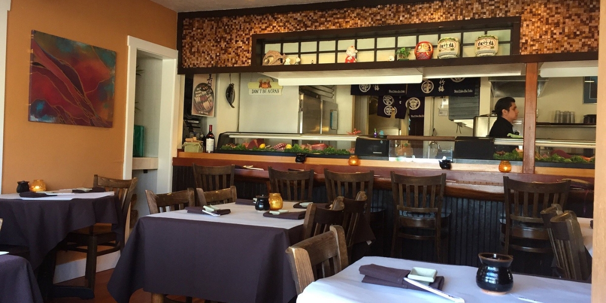 A cozy dining room with linen-covered tables, warm artwork, and an open sushi bar showcasing the inviting interior of Sushi Mambo.
