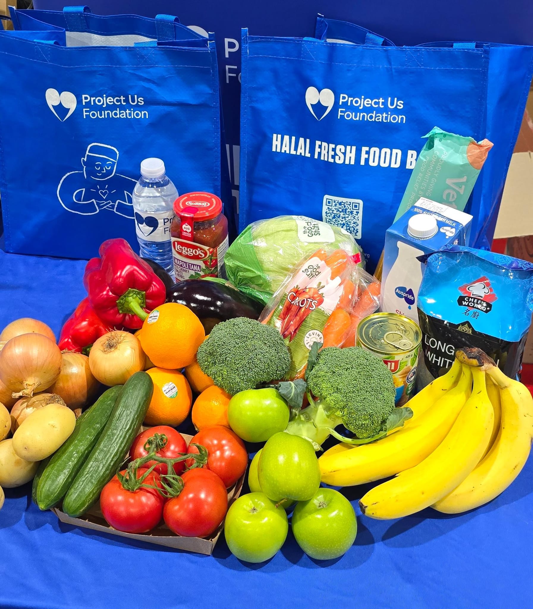 Halal Fresh Food Relief. Variety of fresh groceries including bananas, apples, onions, potatoes, kiwi, lettuce, cauliflower, eggplant, canned chopped tomatoes, chicken cubes, pulse pasta, and a 2-liter milk container, displayed with blue reusable bags branded Project Us Foundation and a banner.