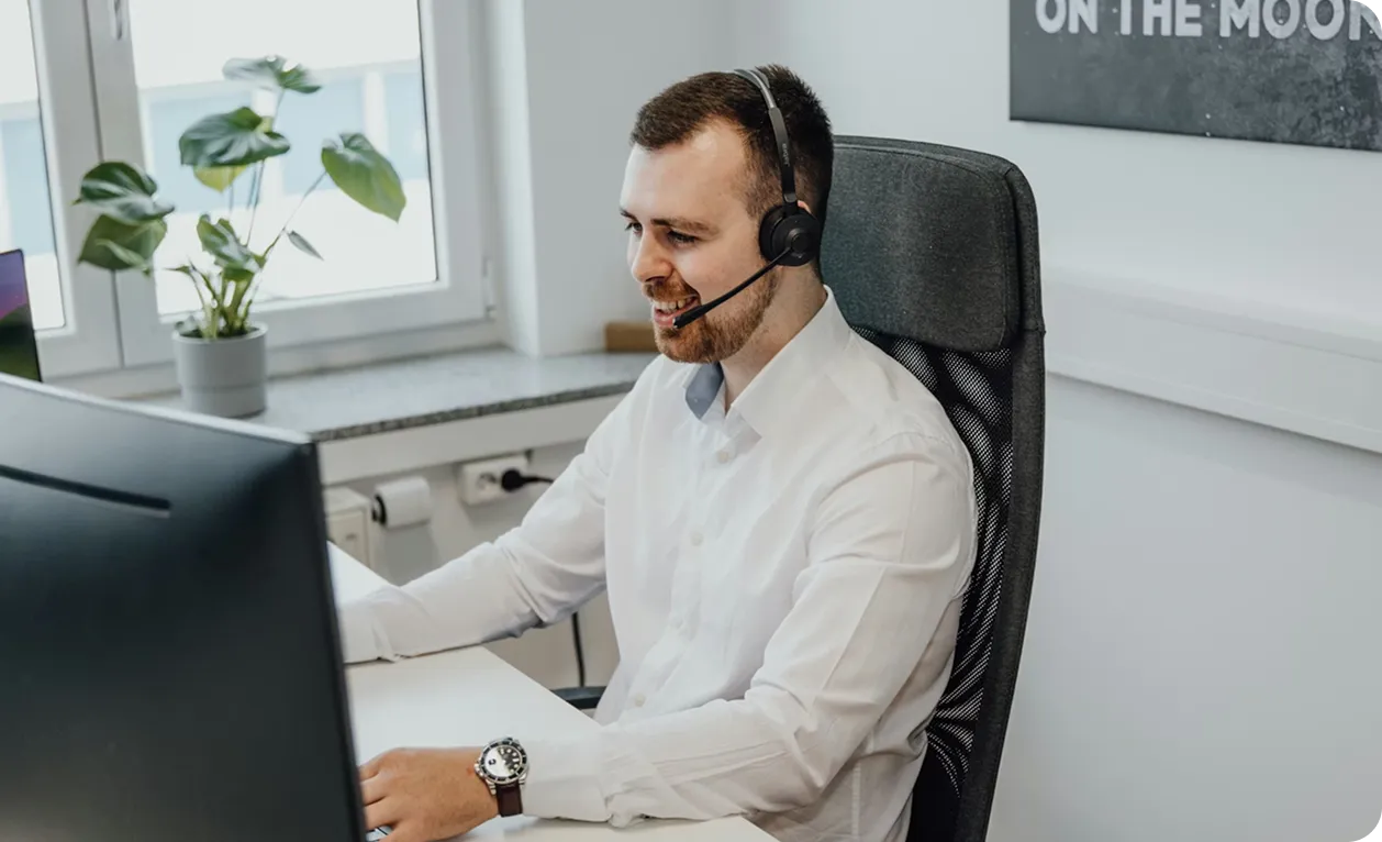 Smiling man wearing a headset and white shirt working at a computer desk in a bright office.
