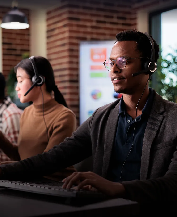 Three customer service representatives wearing headsets working at computers in a modern office with brick walls and large windows.