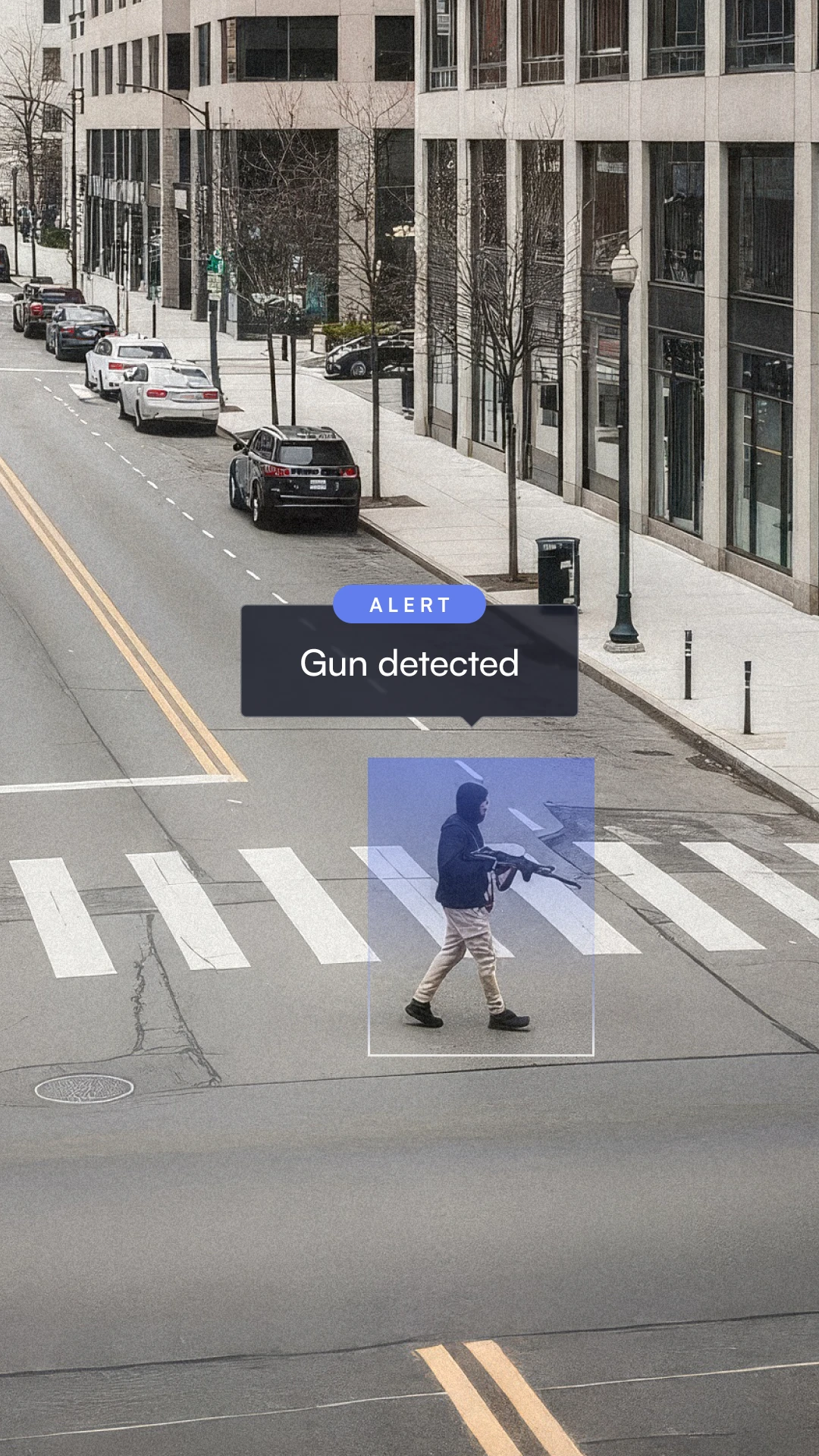 Person wearing a hoodie and light pants walking across a city street carrying a gun, with an alert message reading 'Gun detected.'