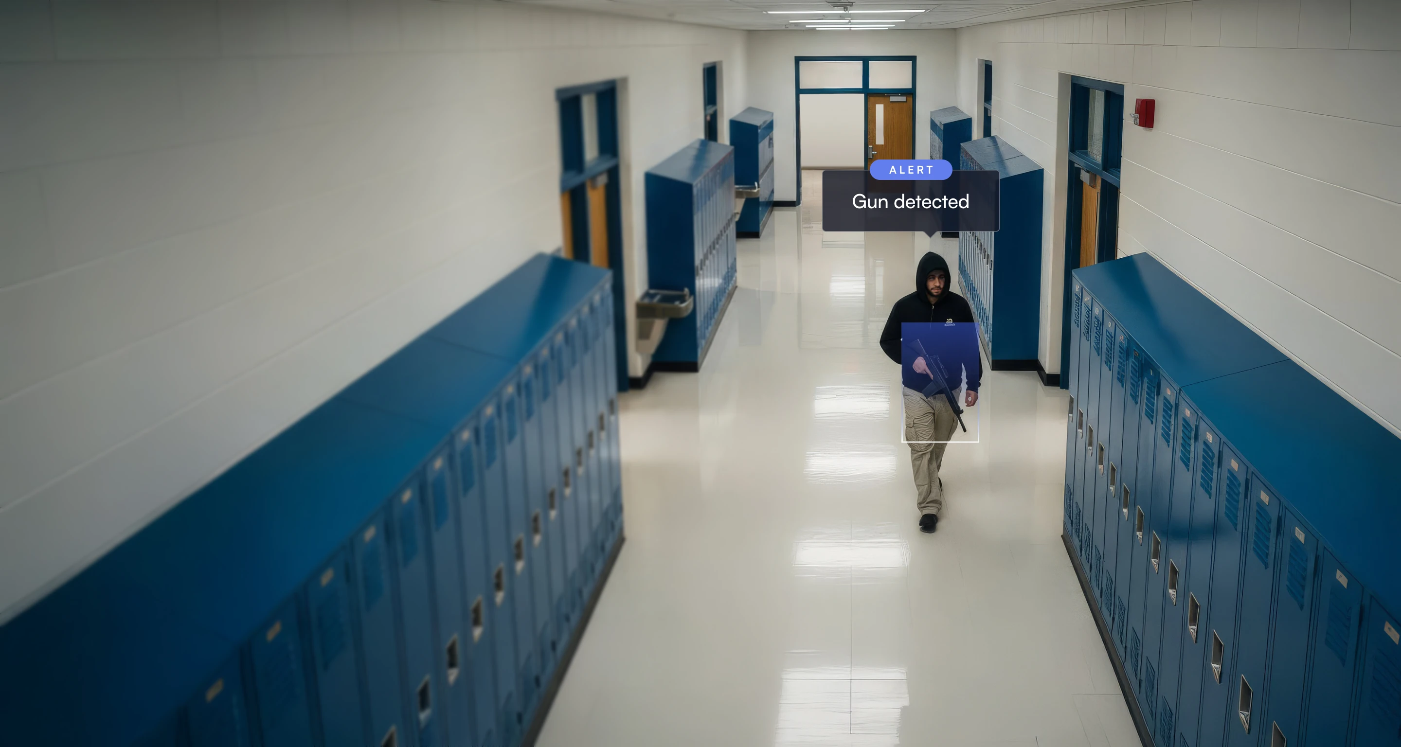 Man wearing a black hoodie walking down a school hallway with blue lockers, holding a gun detected by an alert box.