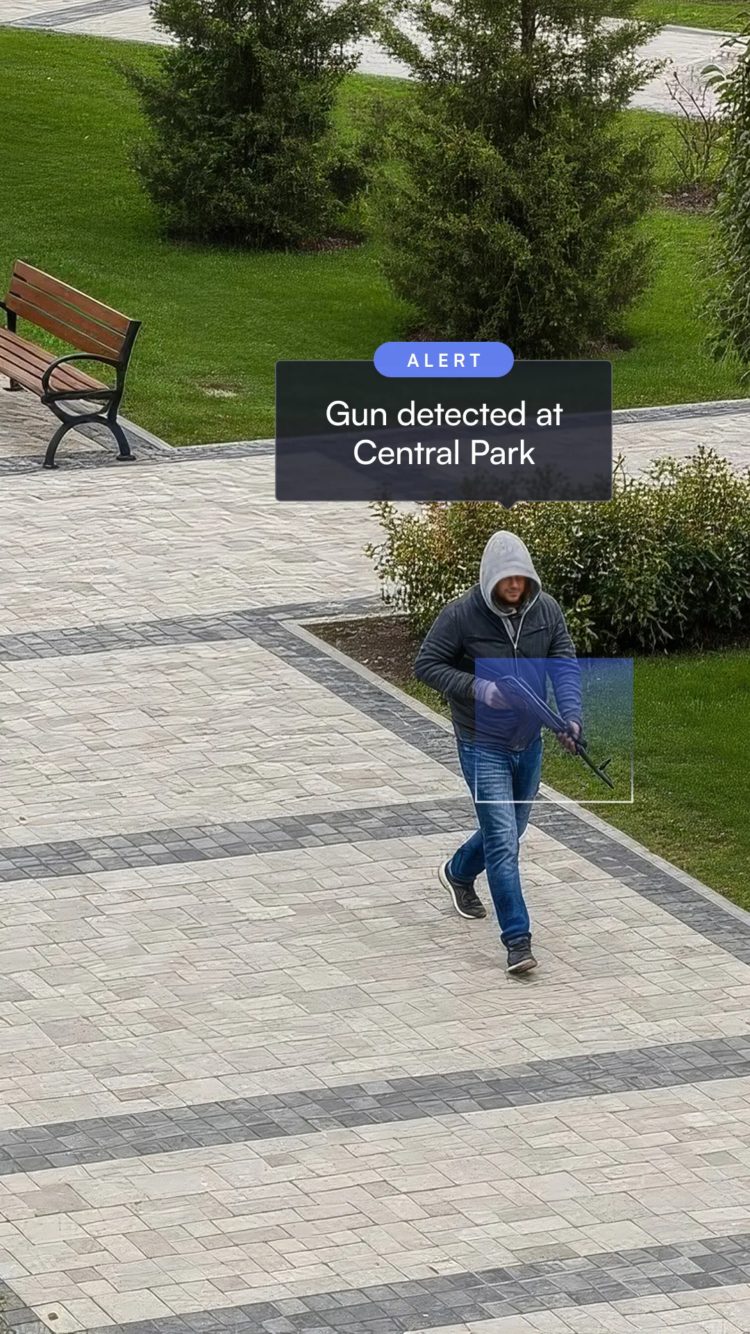 Person in a hoodie walking on a paved park path carrying a rifle, with an alert box saying 'Gun detected at Central Park.'