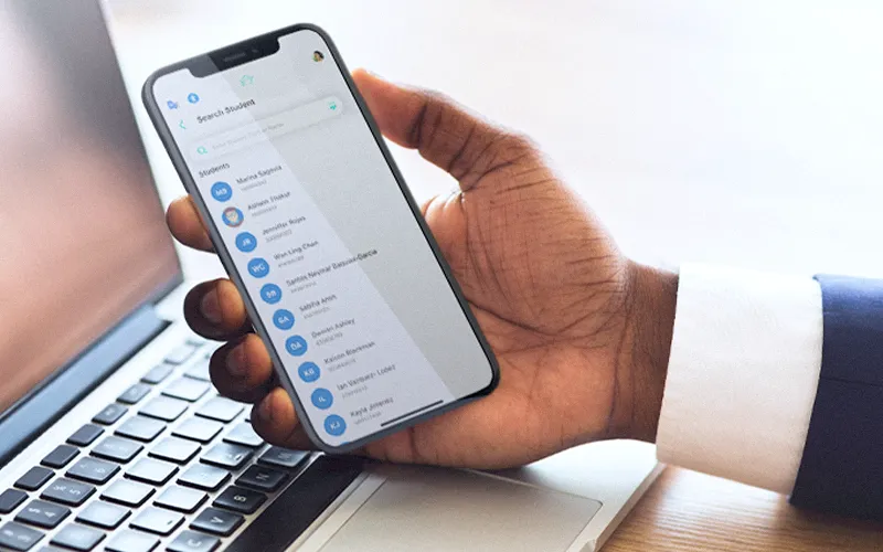 Teacher holding smartphone over laptop showing K-12 STUDENT+ dashboard with grades, attendance and student progress