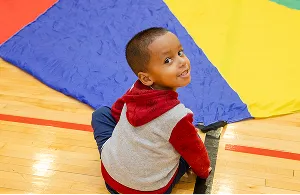 Preschool student smiling while sitting on a colorful parachute during gym playtime