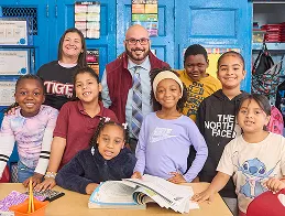 Teacher and a group of elementary students smiling together around a desk in a classroom