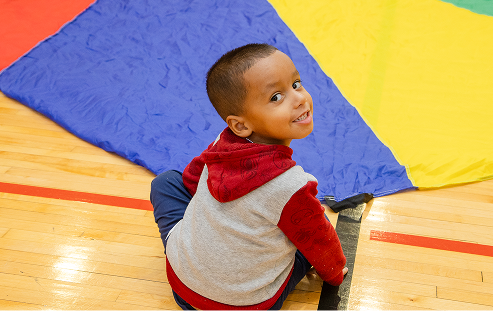 A young child wearing a red and grey hoodie sitting on a wooden gym floor, looking back over their shoulder and smiling near a blue play mat