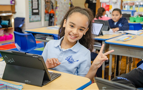 A smiling student sitting at a desk with a tablet computer, holding up a peace sign to the camera