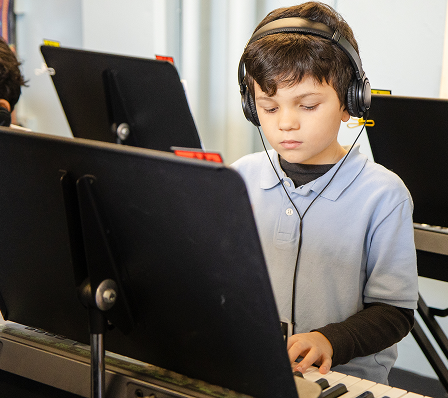 A young boy wearing headphones, looking down with focus at an electronic device or keyboard in a classroom setting.